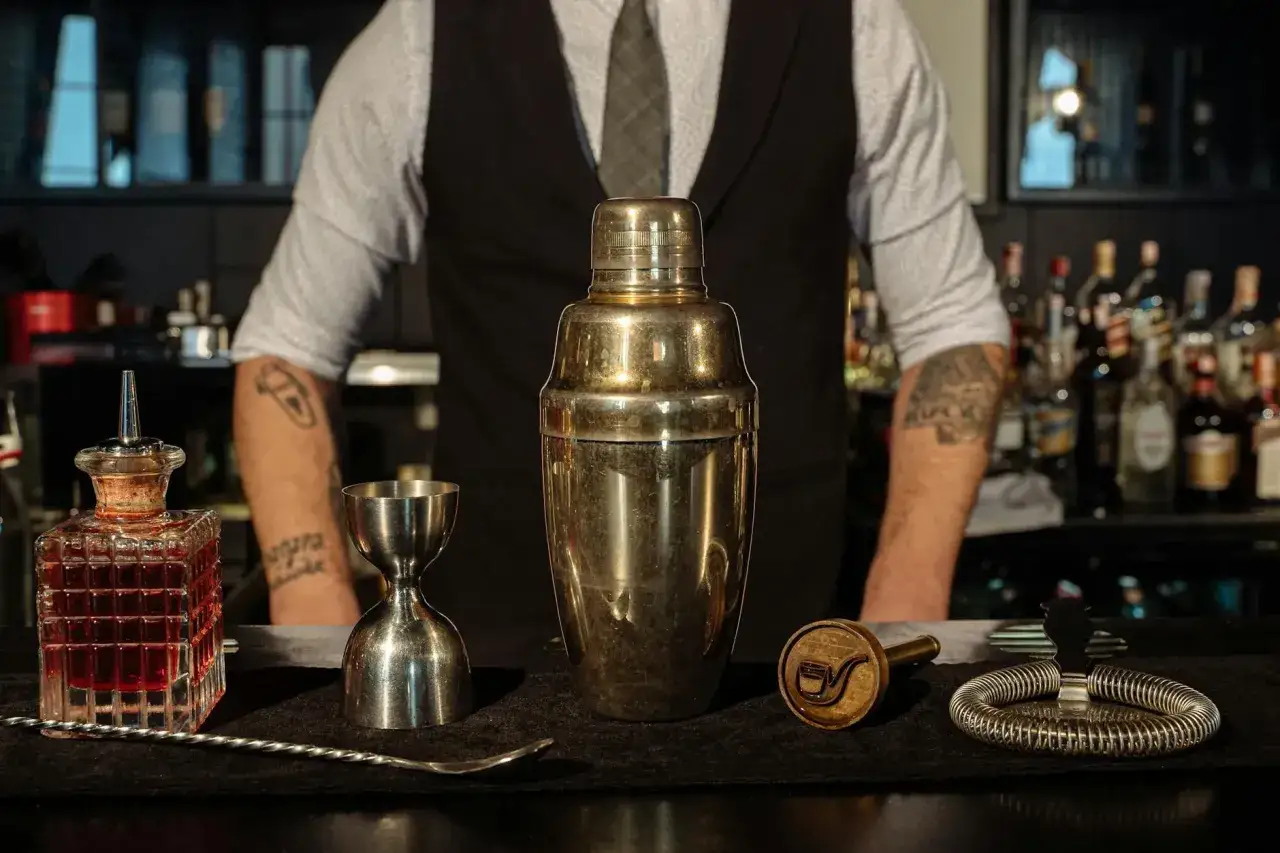 Barman z tatuażami przygotowuje drinka. Na blacie stoją: shaker, jigger, butelka z nalewką, łyżka barmańska, stempel i sitko. Mogą to być przyczyny mętnienia drinków typu stirred przy zbyt długim mieszaniu.