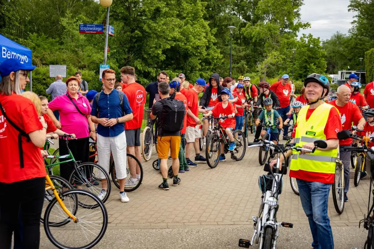 Uczestnicy rajdu rowerowego w czerwonych koszulkach, dzieci i dorośli, gotowi do startu.