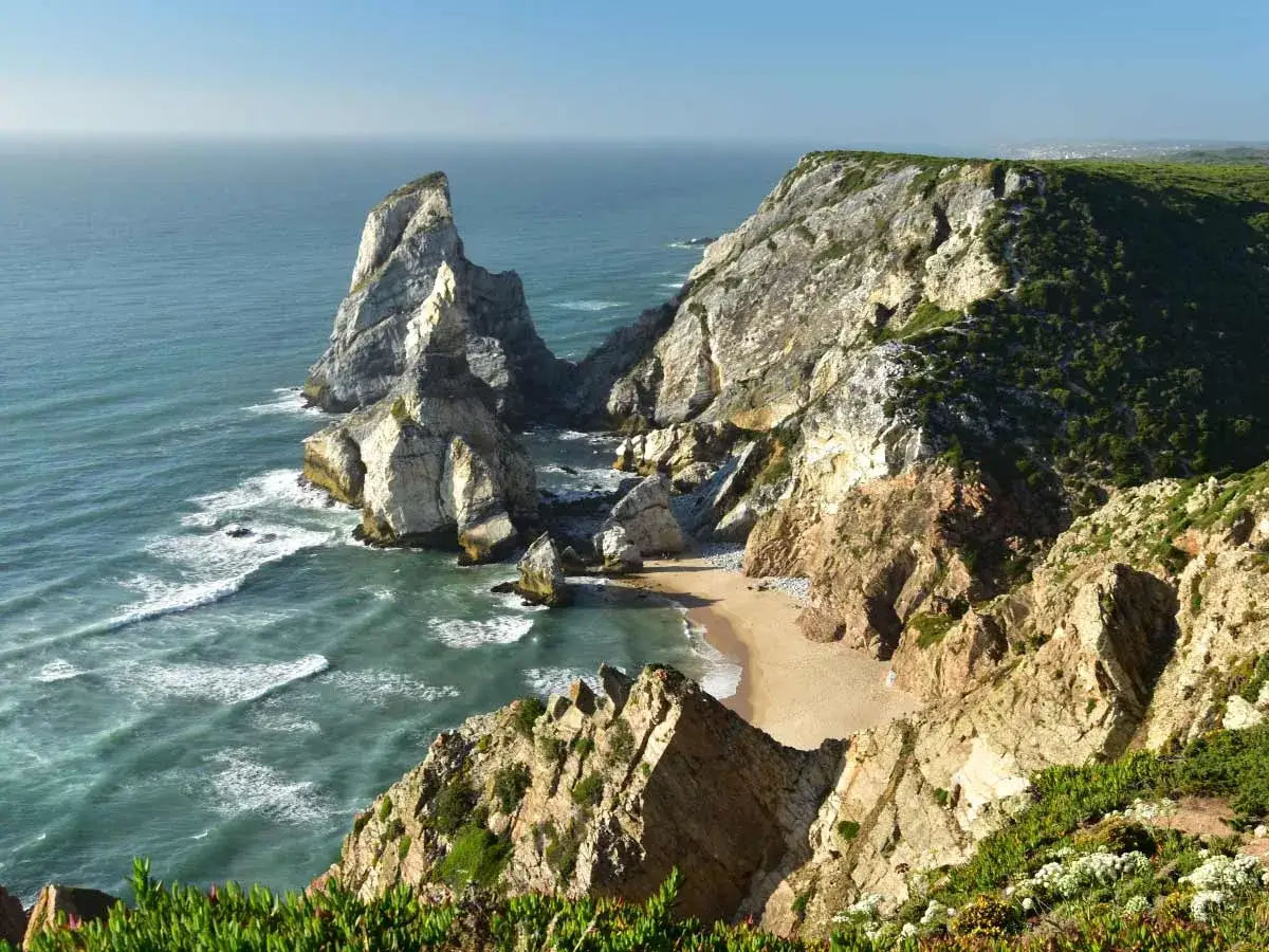 Steile Felsklippen mit &uuml;ppiger Vegetation umgeben einen einsamen Sintra Strand. Das t&uuml;rkisfarbene Meer bricht sich an den Felsen.