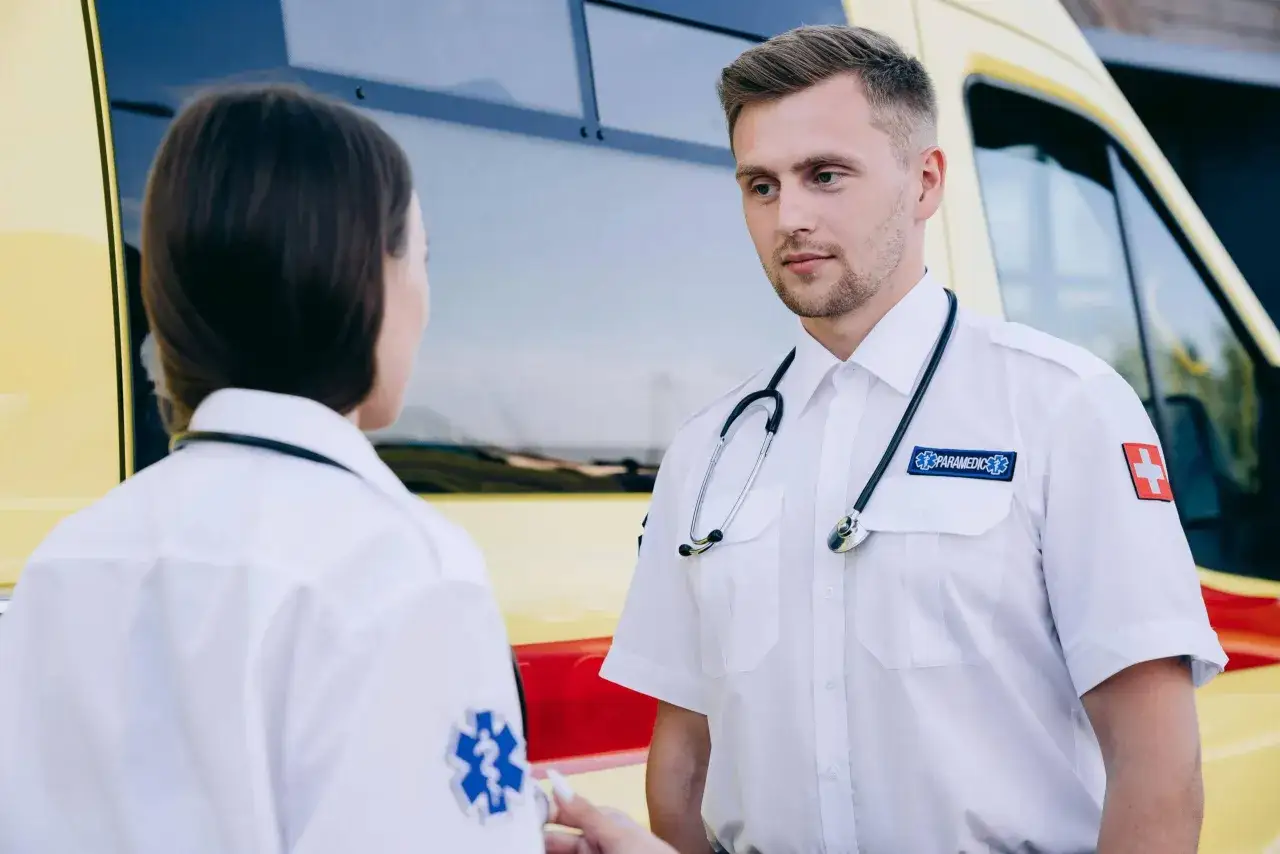Dwóch ratowników medycznych rozmawia przy karetce. Czy studia ratownictwo medyczne są trudne? To wyzwanie, ale warto.