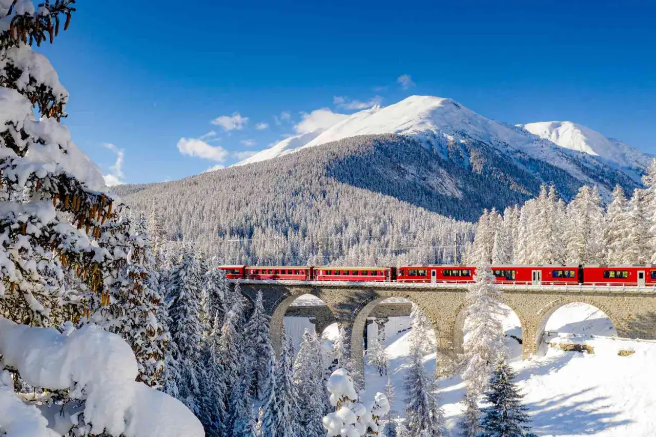 A red train crosses a snowy viaduct, a picturesque journey from St. Moritz to Innsbruck through snow-covered pines and majestic mountains.