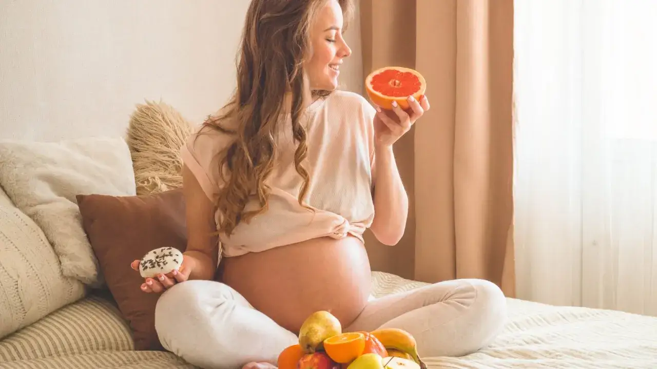 Schwanger, sie hält eine Grapefruit und einen Krapfen, daneben eine Obstschale.