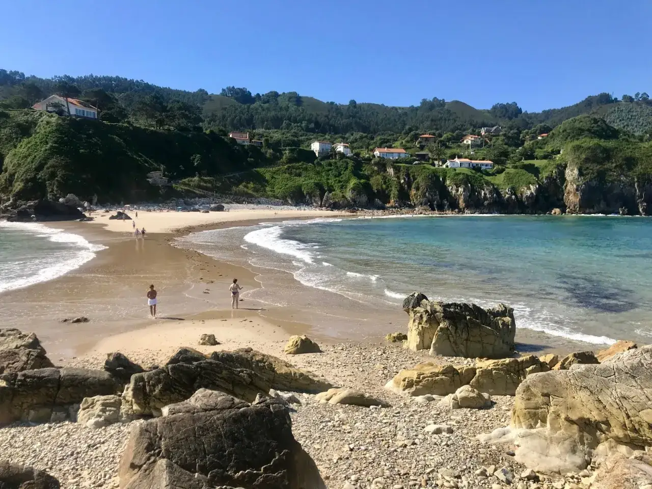 La **playa de Amio** se despliega con arena dorada y aguas turquesas. Dos personas pasean tranquilamente mientras las olas acarician la costa.