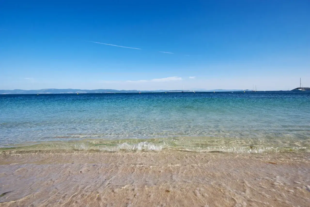 Aguas cristalinas y arena dorada en la playa milide, con veleros navegando en el horizonte bajo un cielo azul intenso.
