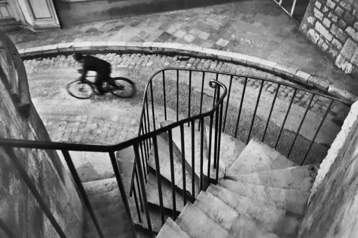 Ciclista sfocato su strada acciottolata, un'immagine che ricorda i grandi fotografi famosi. Scala a chiocciola con ringhiera curva in primo piano.