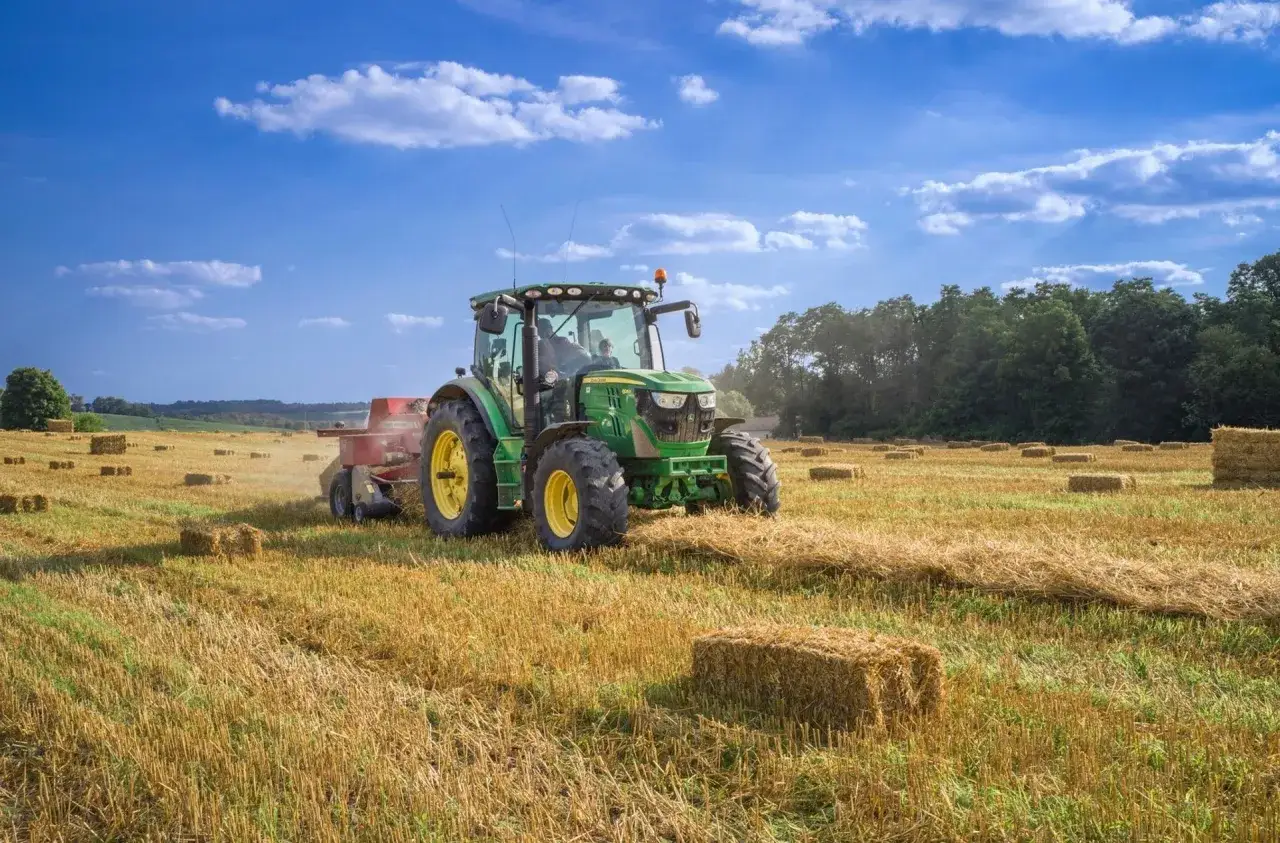 Zielony traktor z prasą zbiera siano na polu. Traktorzysta pracuje w słoneczny dzień, tworząc bele siana.