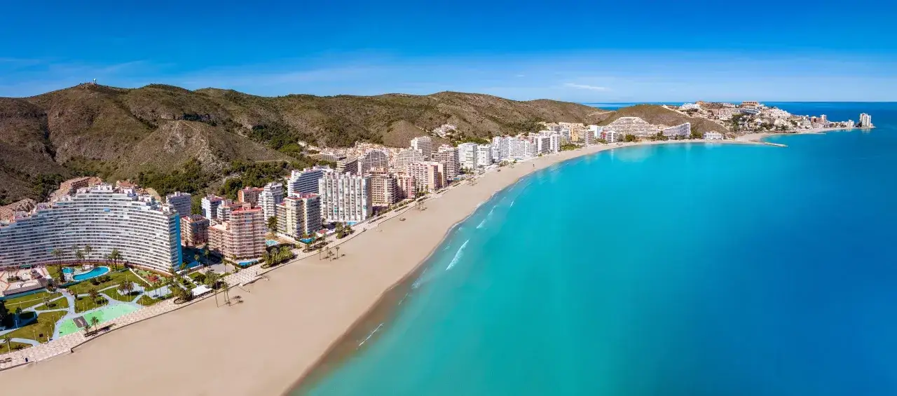 Vista aérea de la costa del balneario Cullera, con edificios residenciales, una playa de arena dorada y el mar turquesa bajo un cielo azul.