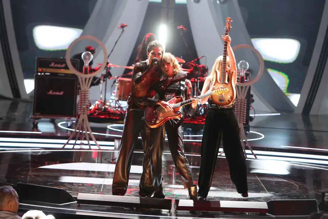 Zespół Måneskin na scenie podczas jednego ze swoich energetycznych koncertów. Wokalista obejmuje gitarzystę, który gra na czerwonej gitarze.