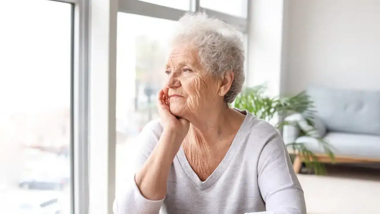 Anziana con mano sul mento, sguardo pensieroso verso la finestra. Potrebbe riflettere su una pseudodemenza depressiva.