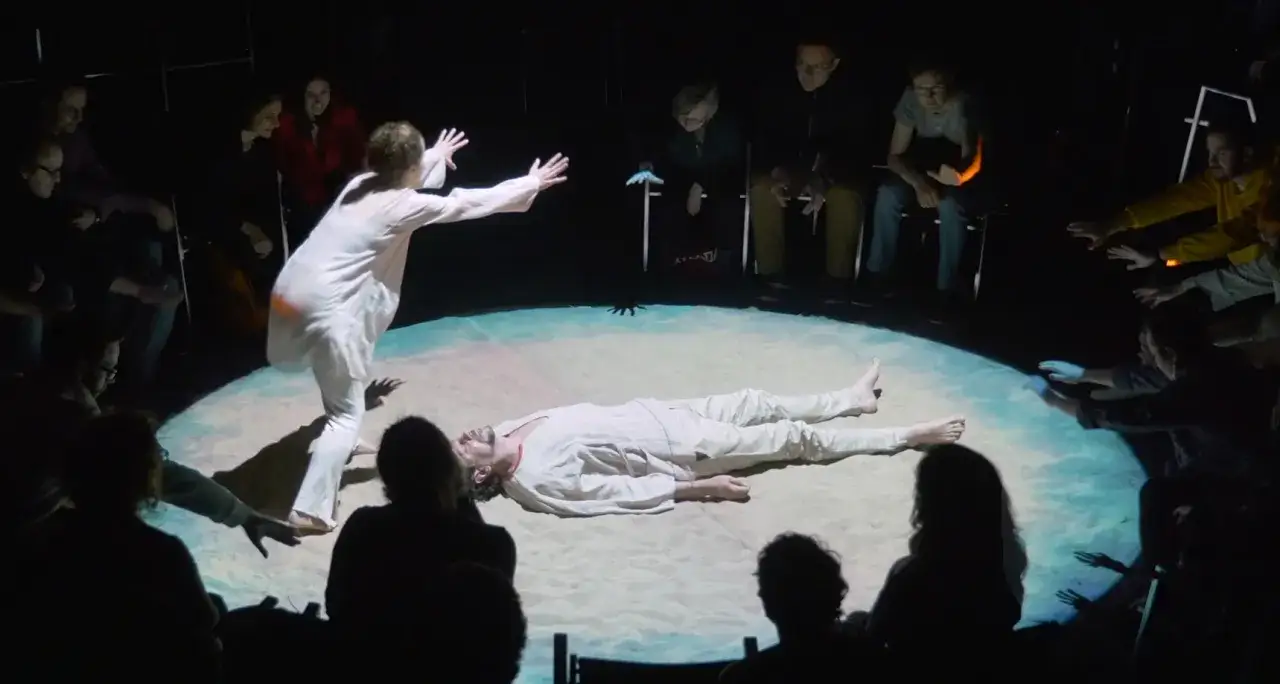 Na scenie sztuki teatralnej leży mężczyzna, a kobieta w białym stroju wyciąga do niego ręce. Widownia otacza scenę, obserwując wydarzenia.