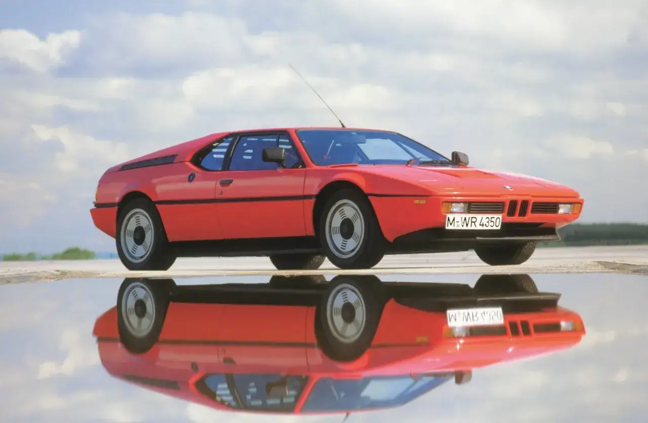 Un ic&oacute;nico m1 coche rojo descansa sobre un espejo de agua, reflejando su silueta deportiva contra un cielo nublado.