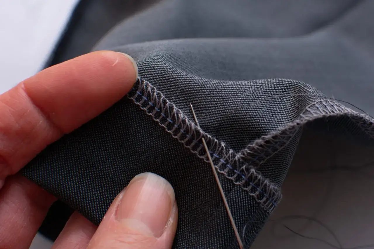 Close-up of a hand demonstrating how to blind hem pants with a needle and thread on dark fabric.