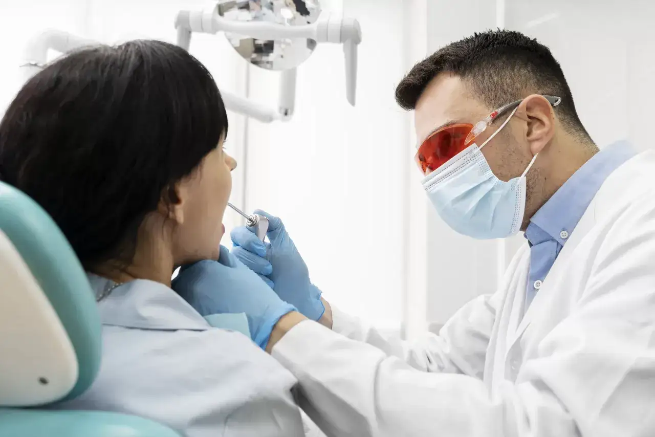 Dentysta w okularach ochronnych i masce bada jamę ustną pacjentki. Rozważane jest migotanie przedsionków a wyrwanie zęba.