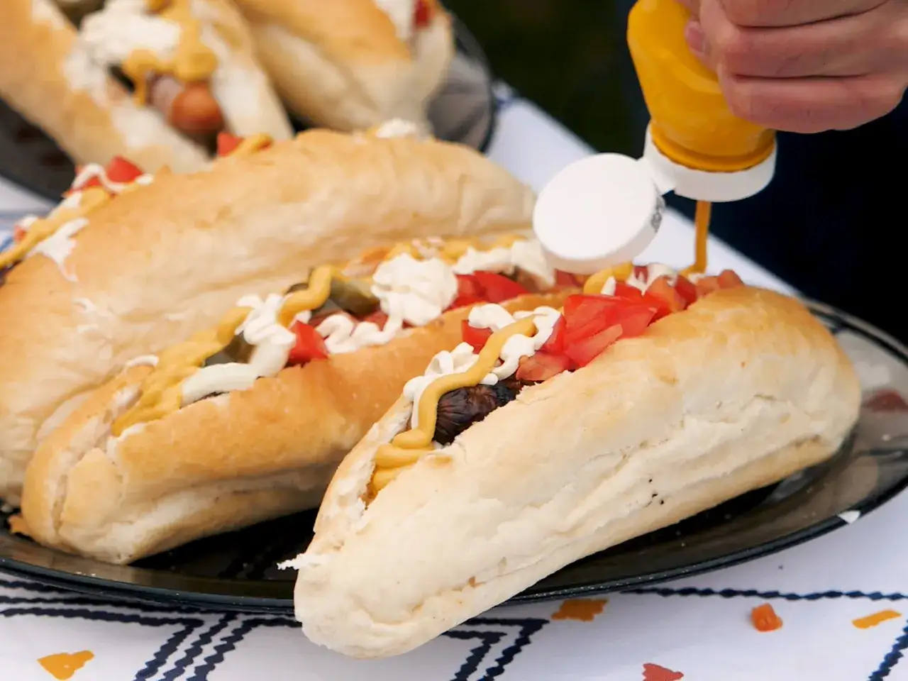 Pyszny hot dog z grilla, polany musztardą, z pomidorami i ogórkiem. Idealny na letnie przyjęcie.