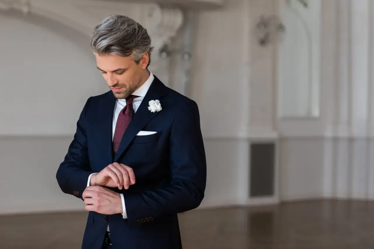 Elegancki mężczyzna w granatowym garniturze, z bordowym krawatem i butonierką, poprawia mankiet. Idealny przykład, jak dobrać garnitur na wesele.