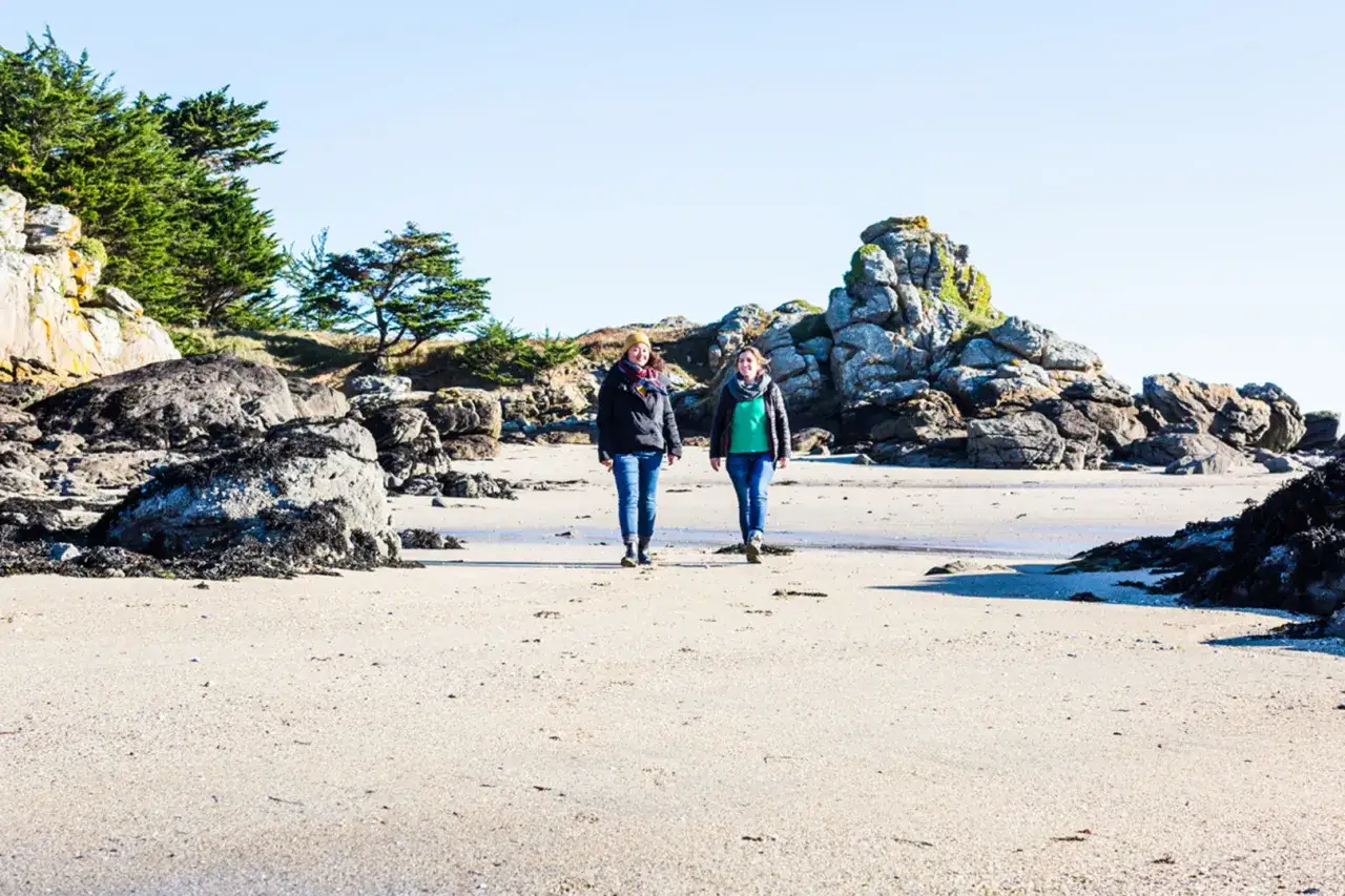 Deux amies profitent d'une randonnée à Saint-Jacut-de-la-Mer, marchant sur une plage rocheuse sous un ciel clair.
