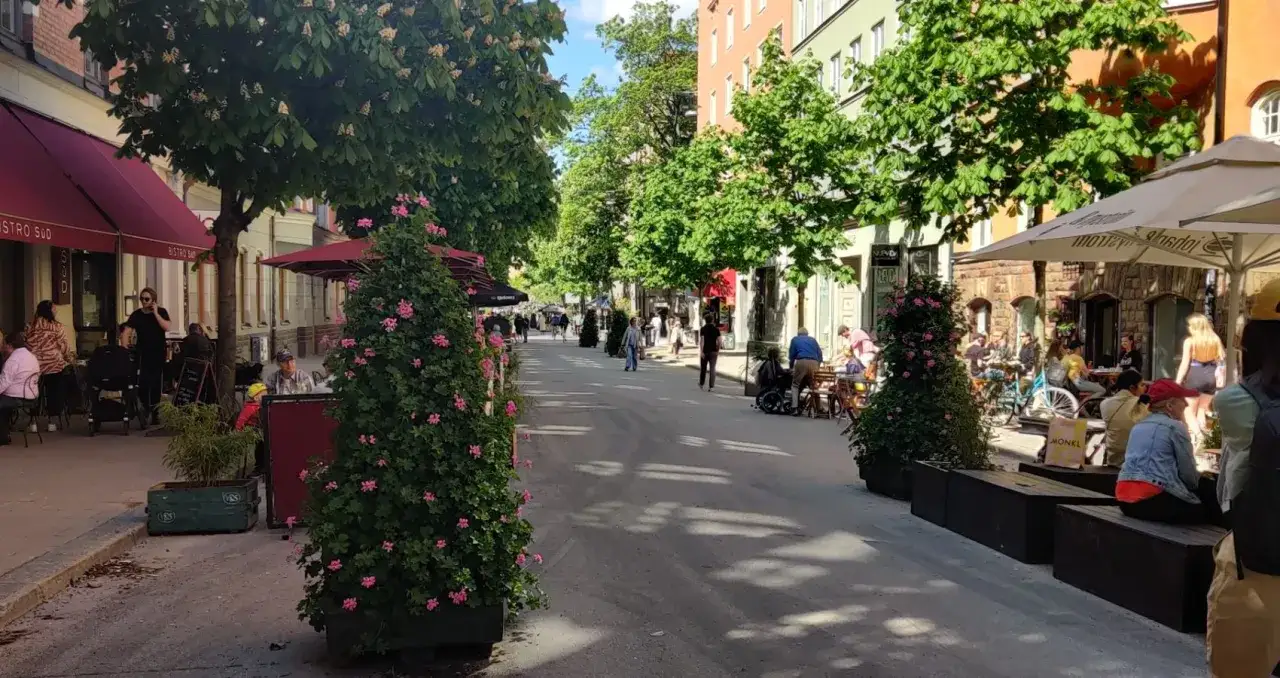 En solig dag i Stockholm, folk njuter av att promenera längs en gata kantad av träd och uteserveringar.