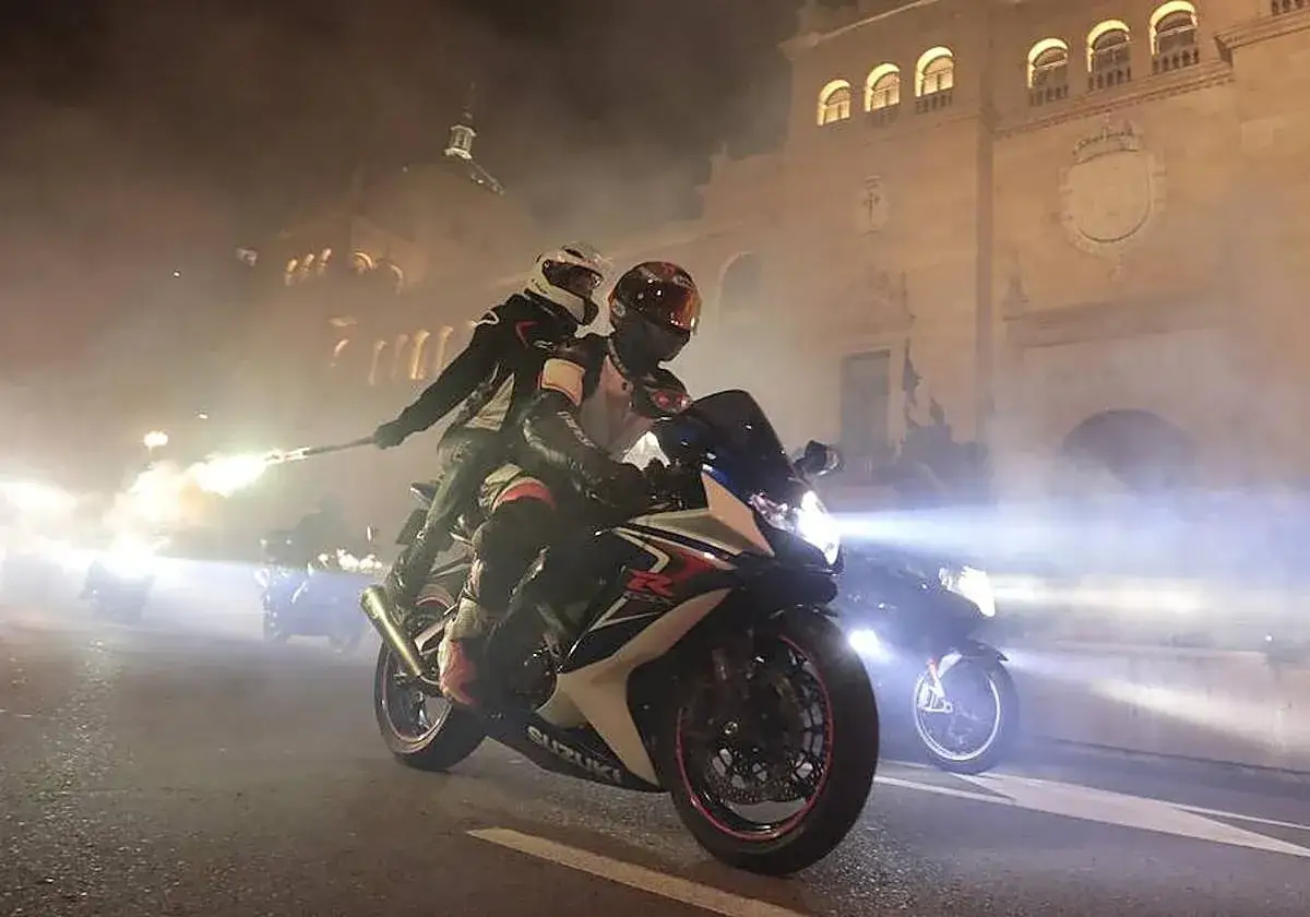 Dos motoristas con cascos circulan de noche, uno lleva una bengala encendida. Una motocicleta puede circular sin tubo de escape, creando un espectáculo de luces y humo.
