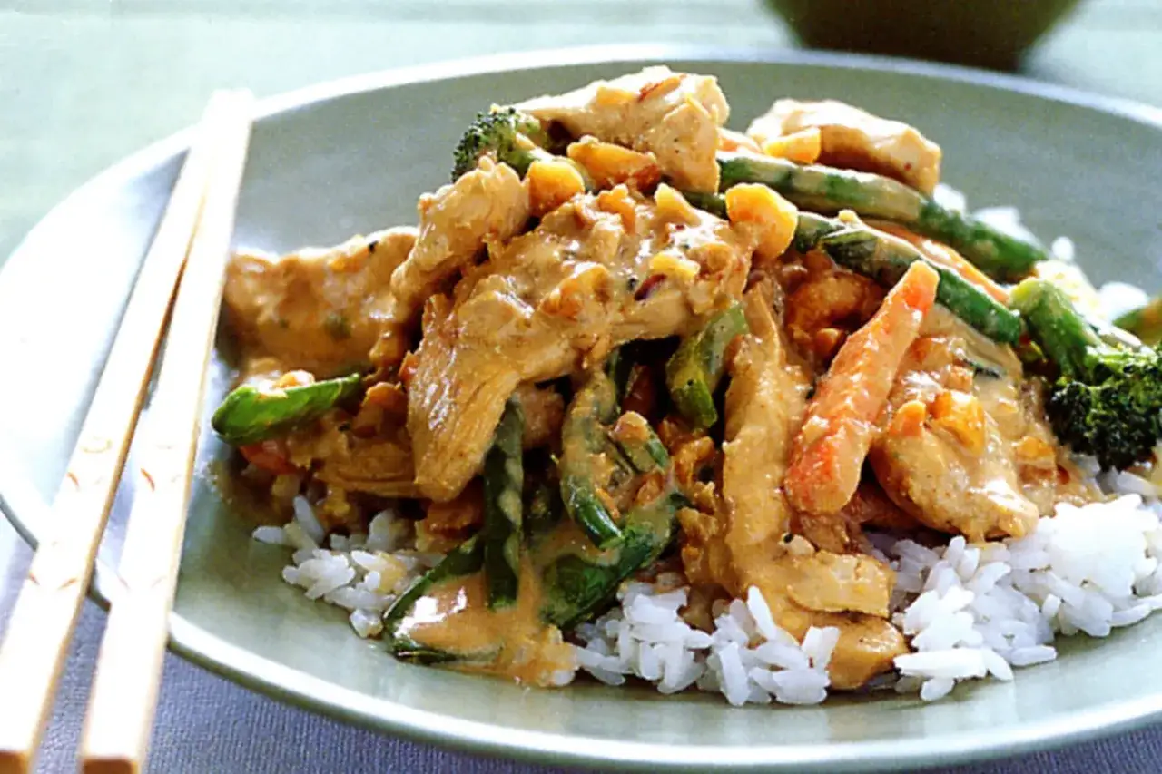Ein Teller mit Reis, bedeckt mit einem cremigen, roten Thai Curry mit H&uuml;hnchen, Brokkoli und Karotten. St&auml;bchen liegen daneben.
