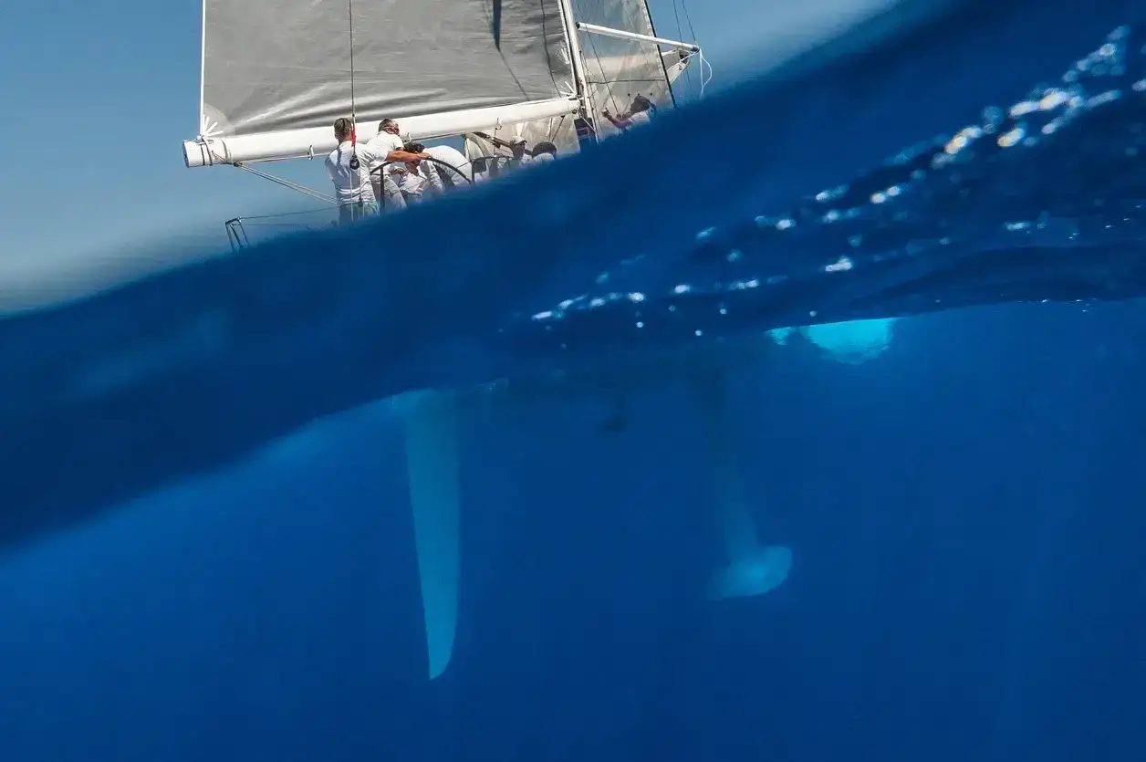 Unterwasseraufnahme des **segelboot rumpf** mit Crew an Deck. Kiel und Ruderblatt ragen in das klare blaue Wasser.