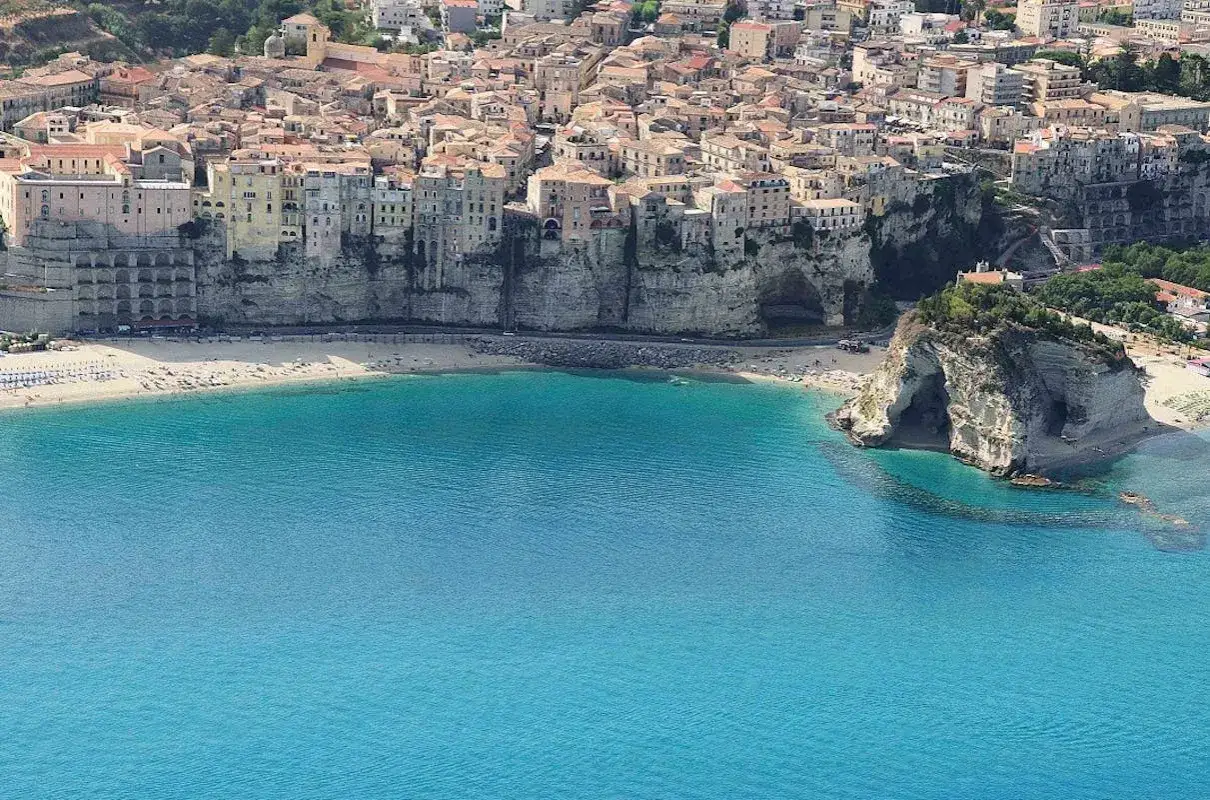 Vista aerea di Tropea, con le case aggrappate alla scogliera e il mare turchese che lambisce la spiaggia. Si intravede il promontorio con le grotte, parte del fascino del castello di Tropea.