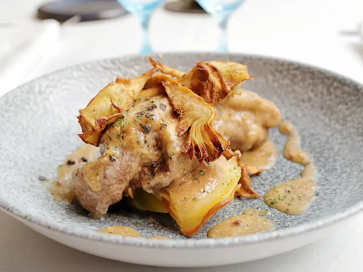 Delicioso ternasco a la pastora, tierno cordero bañado en salsa cremosa y coronado con crujientes alcachofas fritas.