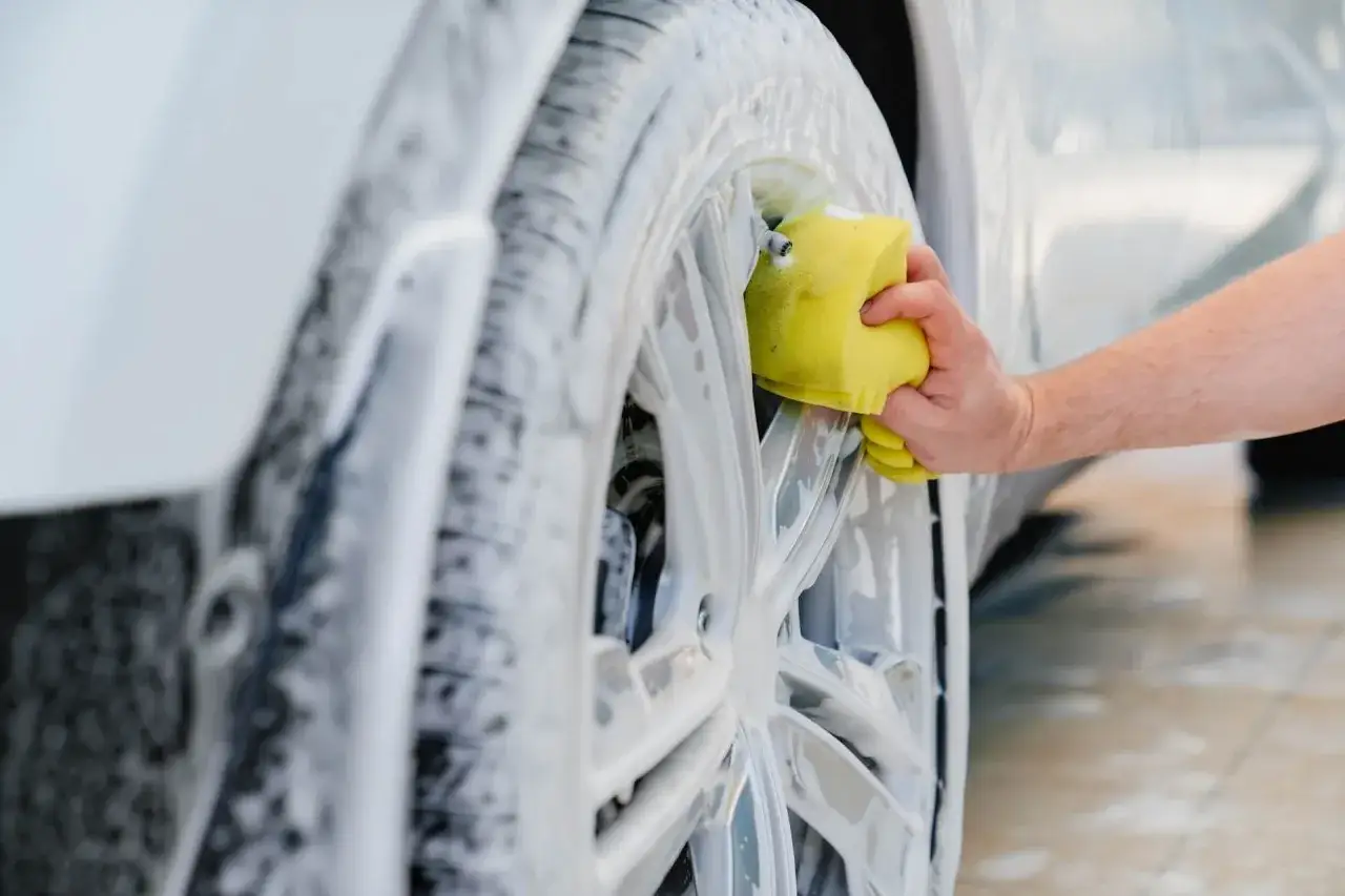 Mano con esponja amarilla limpiando llantas de coche cubiertas de espuma. Un paso esencial para saber como limpiar las llantas del coche.