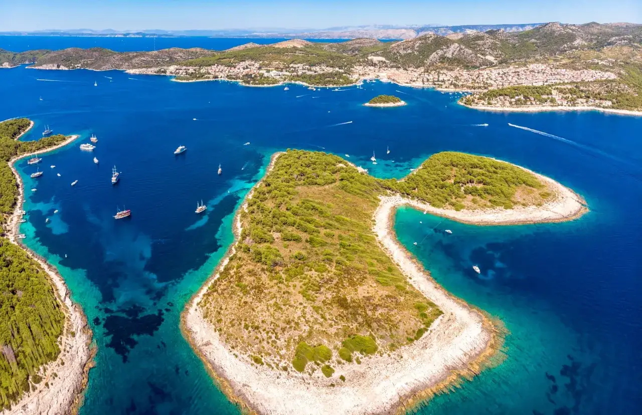 Malownicze Hvar atrakcje: turkusowe morze, zielone wyspy i białe plaże. Wiele łodzi cumuje w zatoce, a w oddali widać miasto.