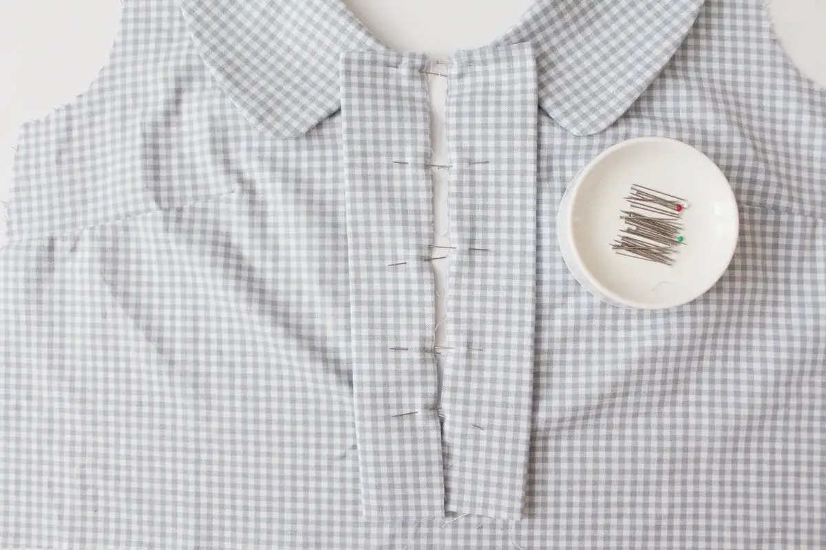 A light blue and white gingham fabric is pinned along the button placket, ready for sewing. A small dish of pins sits nearby.