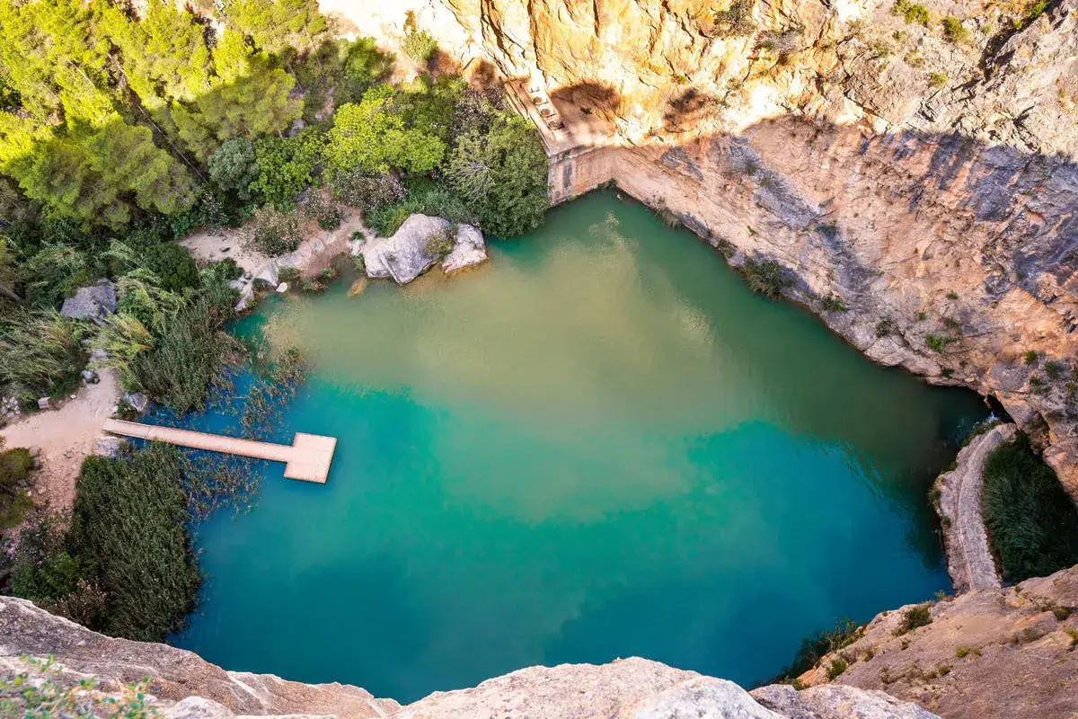 Un **charco azul** en Chiva, rodeado de rocas y vegetación. Un muelle de madera se adentra en sus aguas cristalinas.