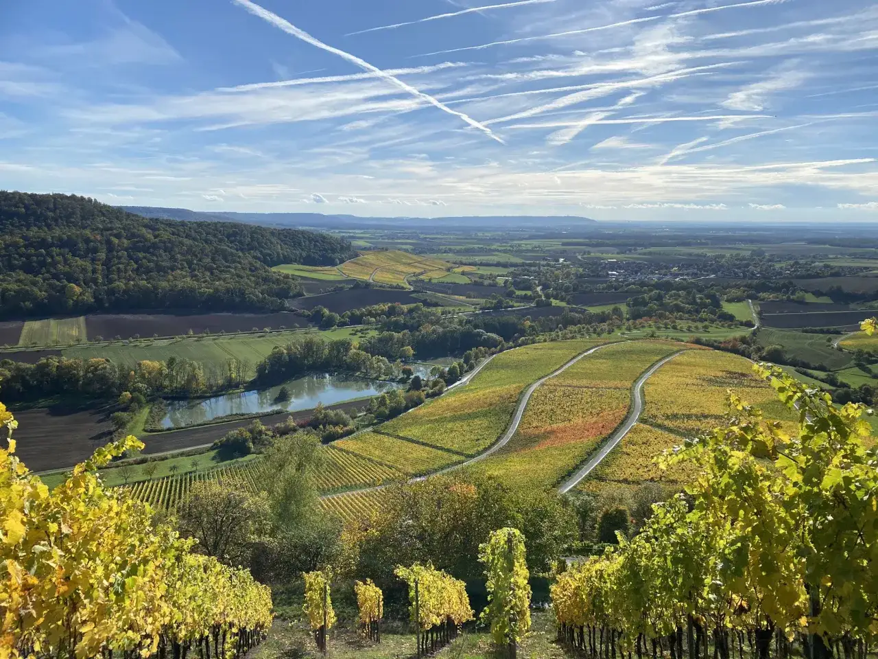 Herbstliche Weinberge im Steigerwald laden zum Wandern ein. Bunte Blätter, ein kleiner See und weite Felder unter blauem Himmel.
