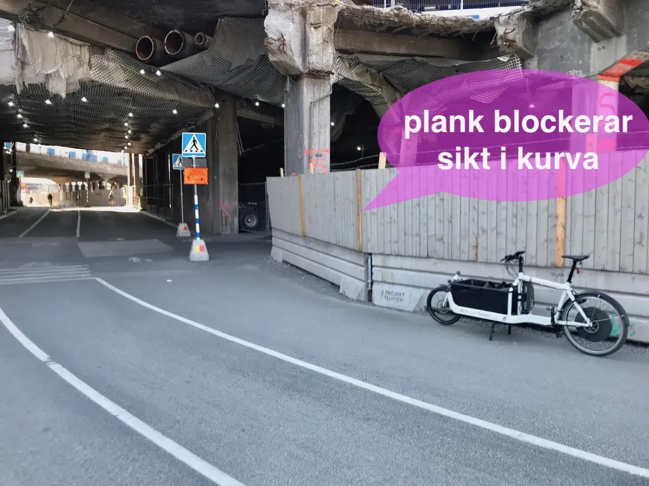 En lastcykel står parkerad vid en väg under en bro. Plankor blockerar sikt i kurvan, vilket skapar en känsla av dold fara.