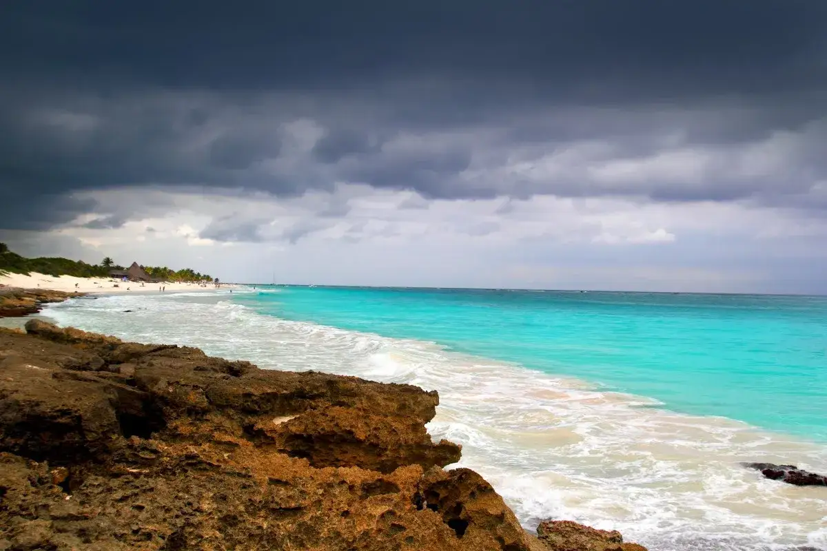 St&uuml;rmische Wolken &uuml;ber dem t&uuml;rkisfarbenen Meer. Ein Strand mit Palmen und Menschen. Meine **Mexiko im Juli Erfahrungen** waren trotz des Wetters unvergesslich.