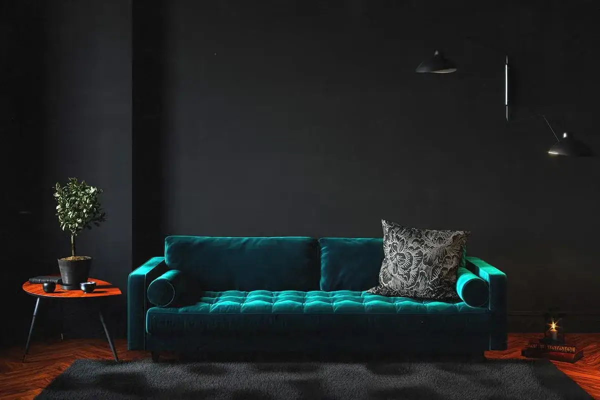 Divano in velluto verde petrolio, con cuscino decorativo, accanto a un tavolino con pianta e lampade a parete.