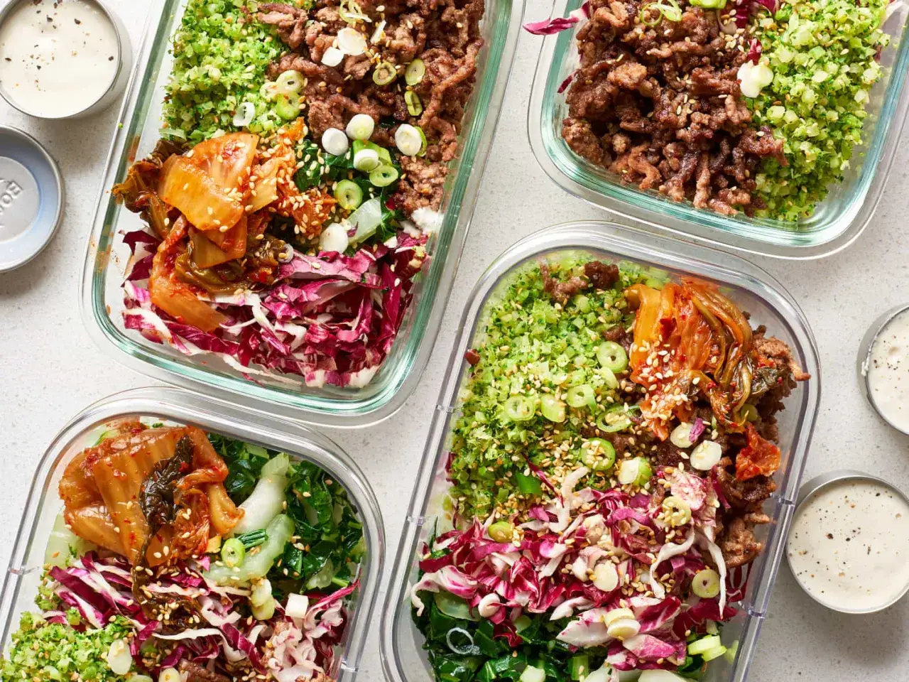 Vier Behälter mit leckeren slow carb rezepten: Hackfleisch, Brokkoli-Reis, Kimchi und Rotkohl. Dazu kleine Schälchen mit Dip.