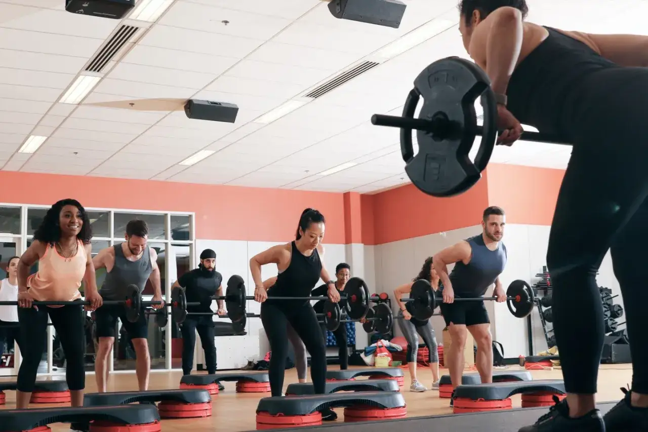 Gruppo in palestra esegue esercizi con bilancieri su step. Il body balance fa dimagrire, tonifica e migliora la postura.