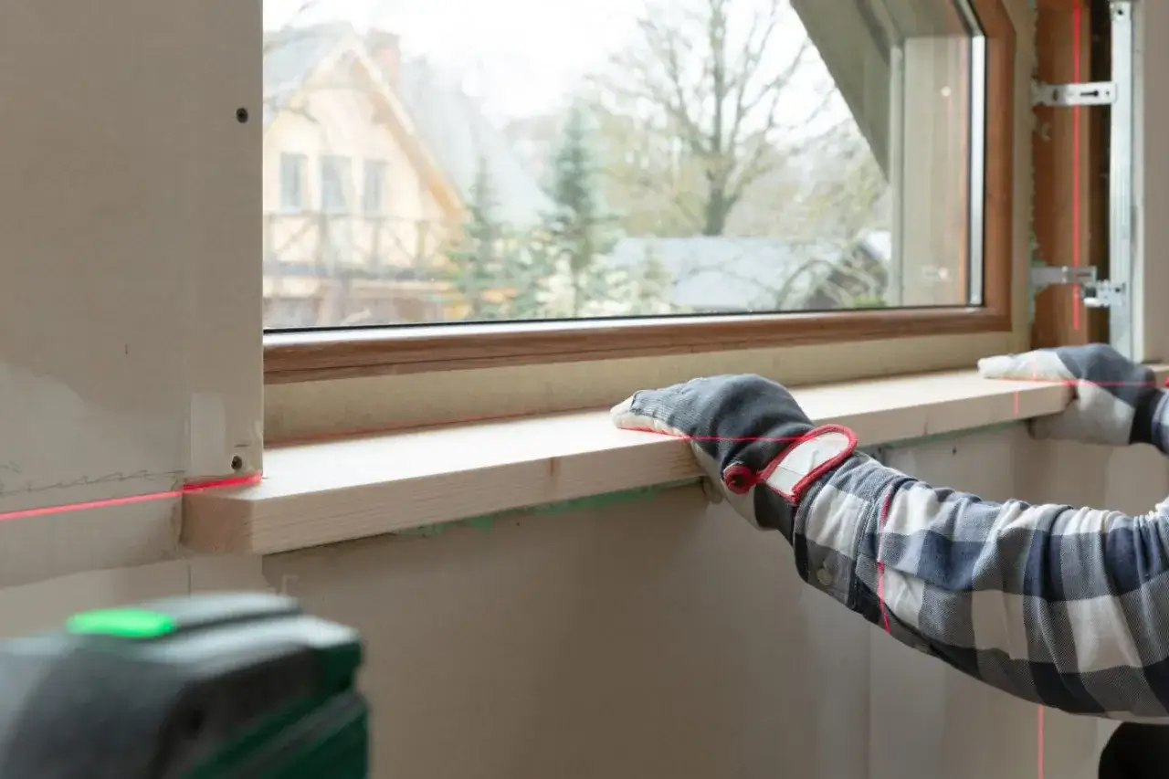 Ręce w rękawicach montują drewniany parapet, używając lasera do precyzyjnego ustawienia. Czy parapet jest konieczny? Tak, dla estetyki i funkcjonalności.