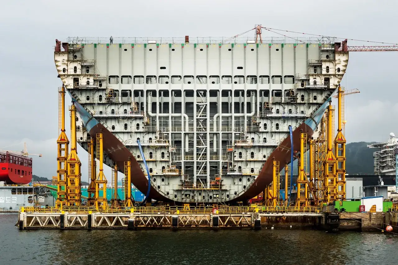 Gigantischer Schiffsrumpf im Bau, gestützt von gelben Gerüsten. Ein Arbeiter auf dem Deck, der die **stowage** (Lagerung/Stauung) im Vergleich zur **storage** (Lagerung/Aufbewahrung) im Schiffbau veranschaulicht.