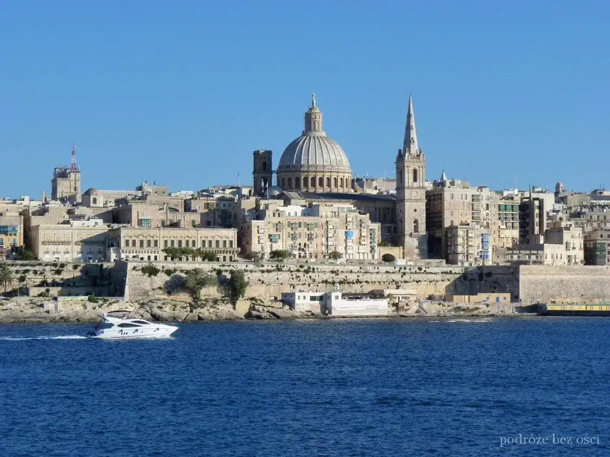 Malta w październiku: biała łódź mija Vallettę z kopułą i wieżą kościelną na tle błękitnego nieba.
