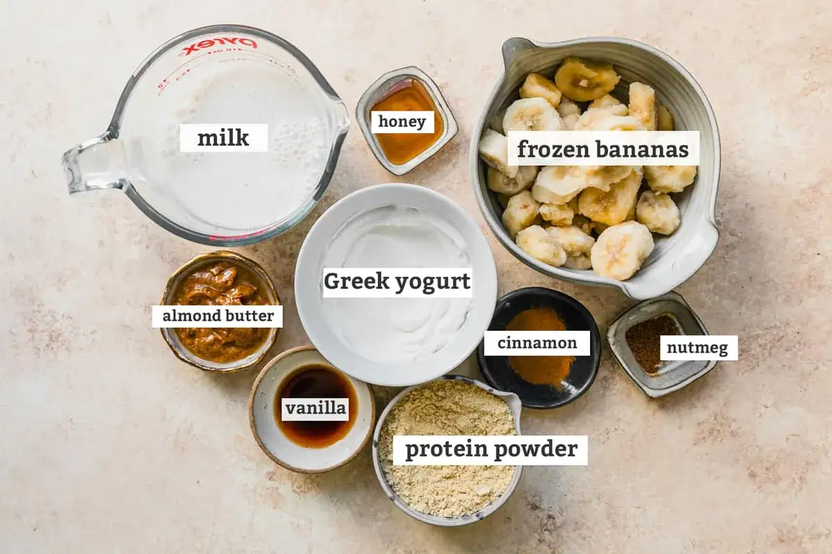 Zutaten für ein leckeres protein shake rezept: Milch, gefrorene Bananen, griechischer Joghurt, Mandelbutter, Honig, Vanille, Zimt und Muskatnuss.