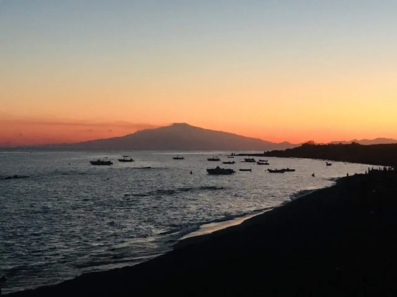 Tramonto sul mare di Reggio Calabria, con barche all'ancora e sagome di montagne all'orizzonte.