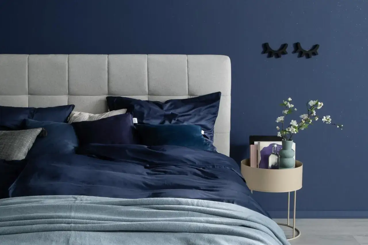 Ein gemütliches Schlafzimmer, das man blau streichen möchte. Dunkelblaue Bettwäsche und ein hellgraues Kopfteil schaffen eine beruhigende Atmosphäre.