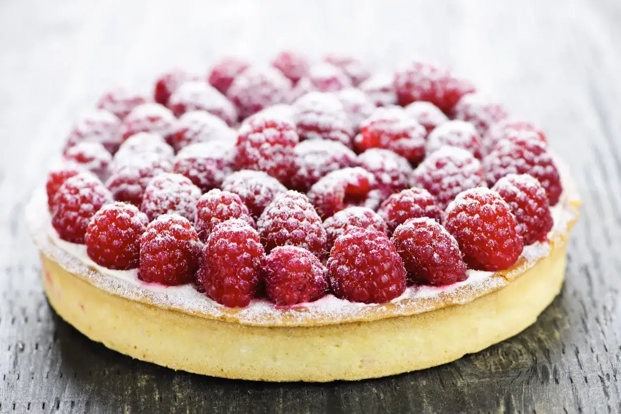 Une tarte à la framboise facile, couverte de sucre glace, sur un fond de bois sombre.