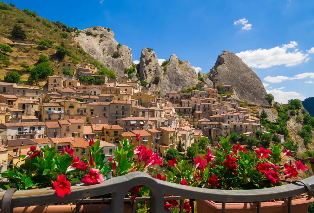 A picturesque view of Pietrapertosa, Italy, a village clinging to a mountainside, with vibrant red flowers in the foreground.