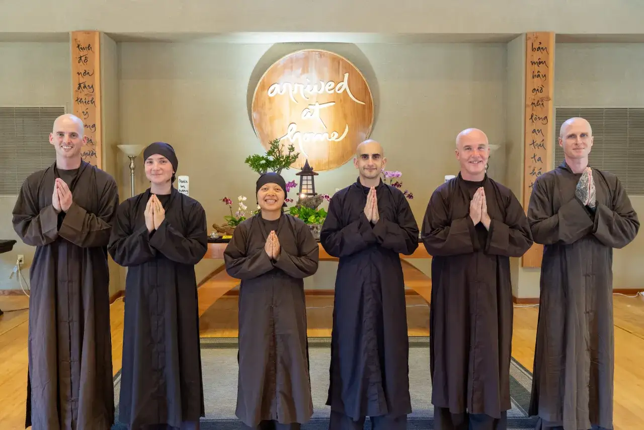 Six monks, including Brother John and Sister Mary, stand with hands pressed in prayer. They are in a serene room with