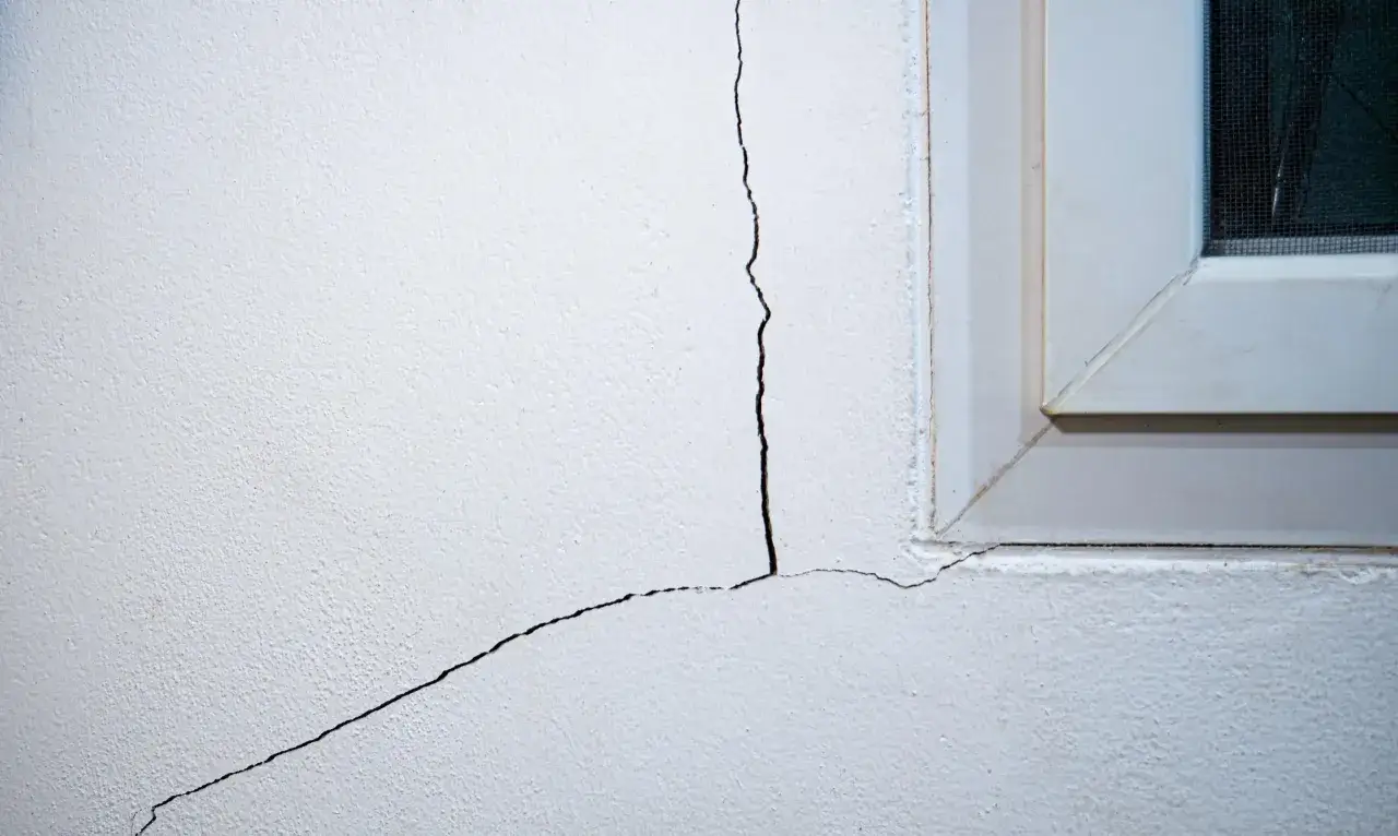 Tipos de grietas en paredes: una grieta fina y otra m&aacute;s gruesa se extienden desde la esquina de una ventana blanca hacia el interior de la pared.