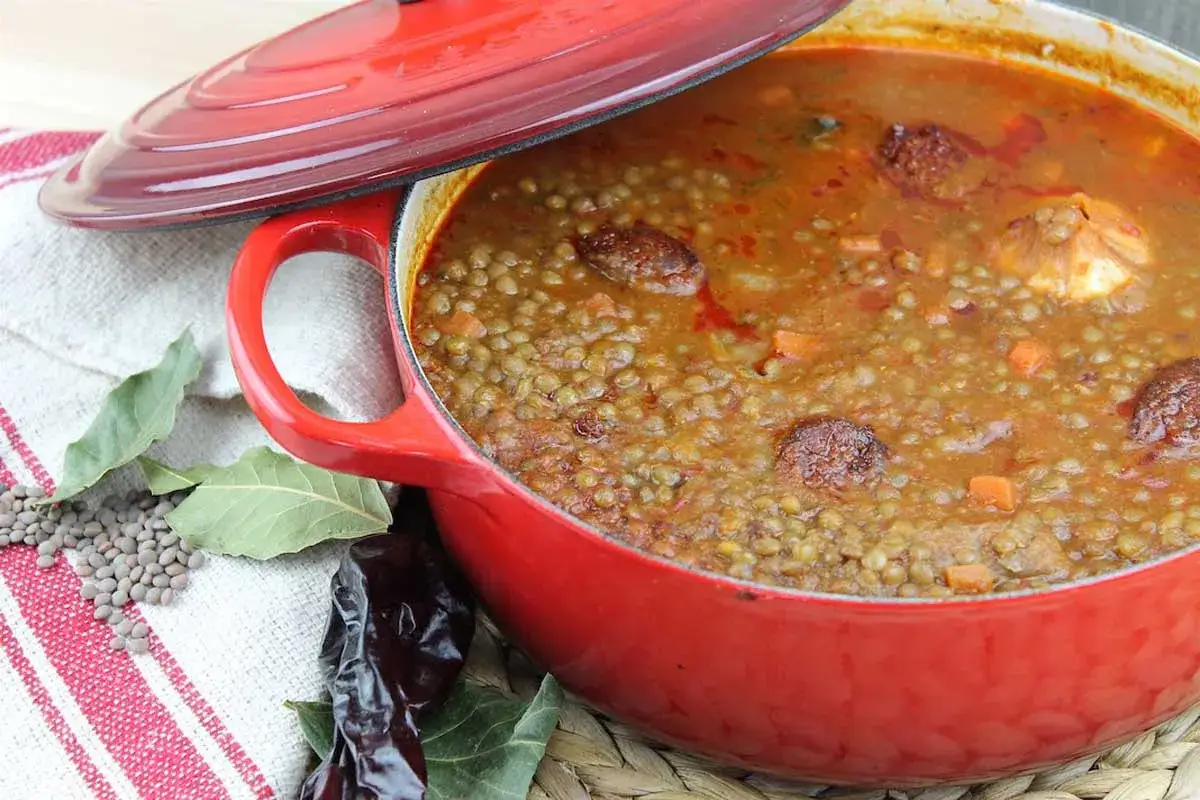 Un reconfortante potaje de lentejas humeante en una olla roja, con trozos de chorizo y verduras.