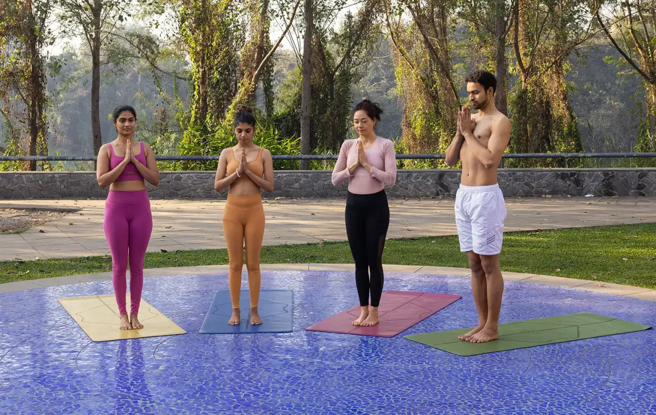 Quatre personnes pratiquent le hatha yoga en extérieur, mains jointes en prière, sur des tapis colorés.