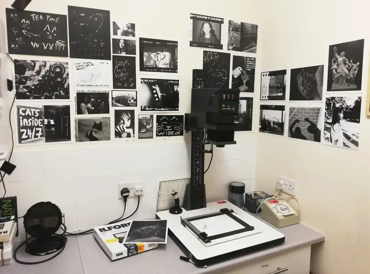 Pracownia fotograficzna, ciemnia z powiększalnikiem, odbitkami na ścianach i materiałami do wywoływania.