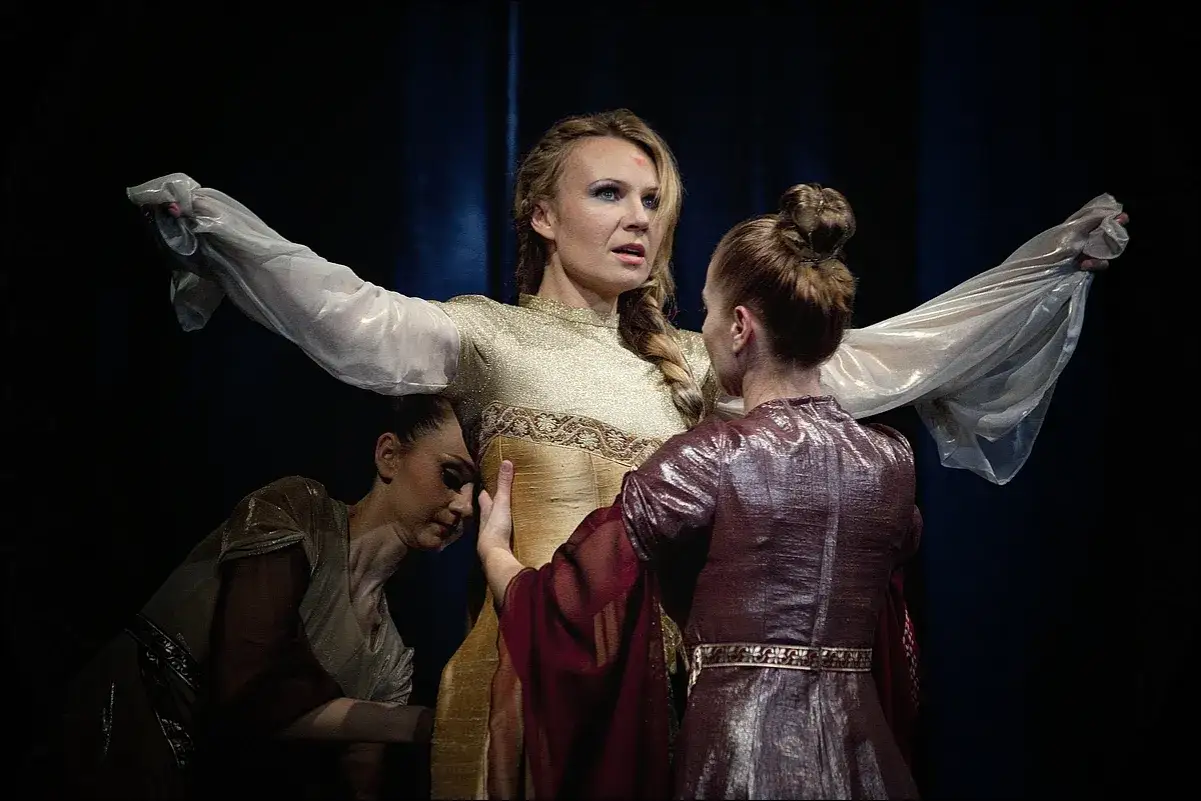 Anna Guzik w złotej sukni, otoczona przez dwie tancerki w teatralnym ujęciu.